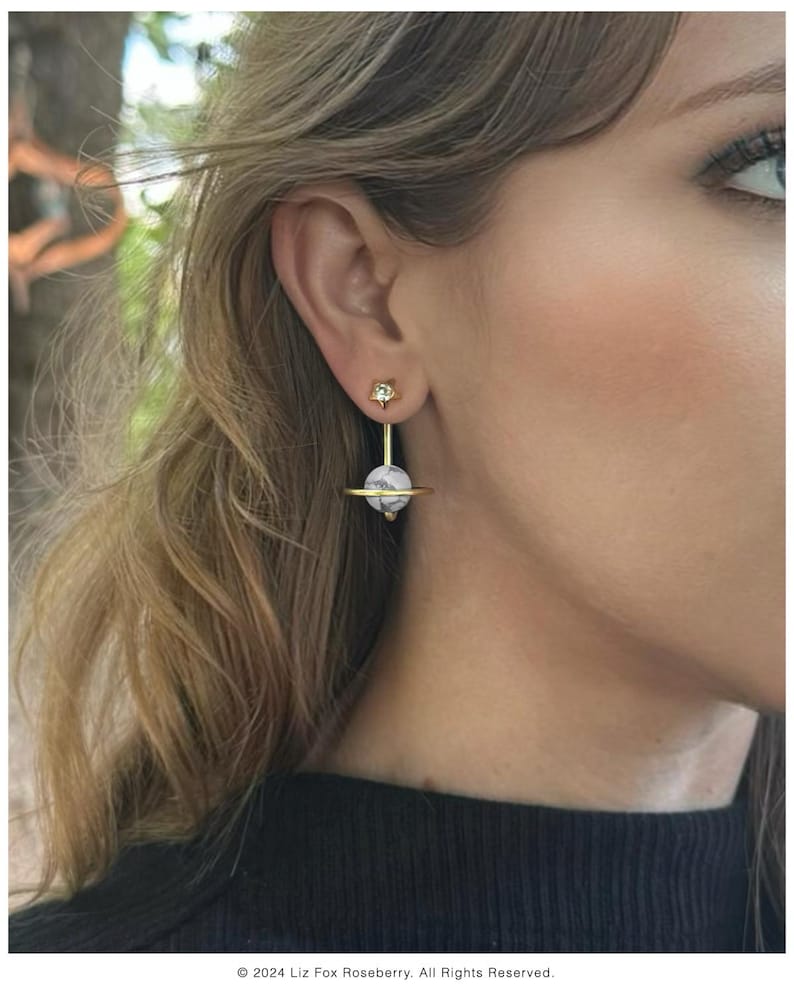 May include: Gold-tone earrings with a celestial design. Each earring features a small star-shaped stud, a vertical gold bar, and a round, marbled stone. A gold ring encircles the stone. The earrings are a unique accessory.