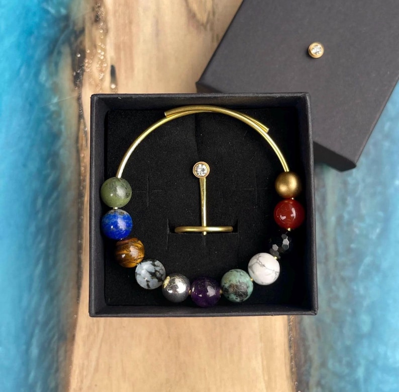 May include: A gold-toned bracelet with colorful gemstone beads, presented in a black box. The bracelet features a variety of stones, including green, blue, and red, and is paired with a matching ring. A small, round, gold-toned stud is on the box lid.