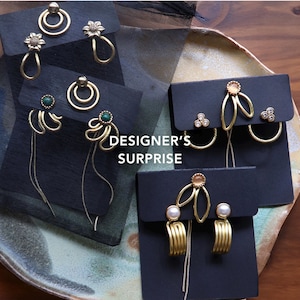 May include: A collection of gold-toned earrings displayed on black cards, featuring various designs including floral motifs, circular hoops, green stone accents, and pearl-like beads. The text "DESIGNER'S SURPRISE" is centered among the earring sets.