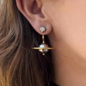 May include: A pair of gold and silver earrings featuring a small round gemstone and a larger sphere with a ring around it, resembling a planet.