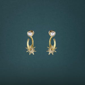 May include: A pair of gold-toned earrings featuring a white spherical pearl stud at the top, connected to a curved gold hoop that suspends a sunburst charm embellished with small clear crystals radiating from a central circular frame.