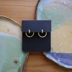 Peut inclure: Une paire de boucles d'oreilles créoles dorées présentées sur une carte noire. Une boucle d'oreille a un motif texturé, l'autre est lisse. Les boucles d'oreilles sont posées sur un fond d'assiettes en céramique vertes, jaunes et bleues.