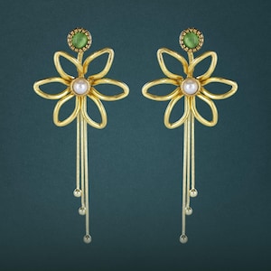 May include: A pair of gold-toned earrings featuring a six-petaled flower design with a central white pearl. Above the flower sits a small green cabochon stone. Three thin gold chains with small spherical beads hang from the base of each flower.