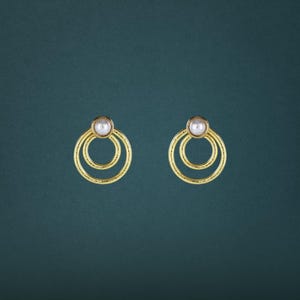 May include: A pair of gold-toned earrings featuring a textured double-circle design. Each earring is accented with a single, smooth, round white pearl centered at the top of the concentric circles, creating a balanced and geometric appearance.