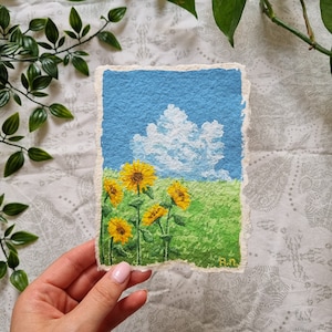 Op de afbeelding: Een handgeschilderd kunstwerk dat een zonnig landschap met zonnebloemen toont. De schildering toont een blauwe lucht met een pluizige witte wolk, een groen veld en gele zonnebloemen. Het kunstwerk is op getextureerd papier.