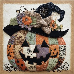 Könnte beinhalten: Ein Patchwork-Halloween-Kürbis mit Hexenhut. Der Kürbis besteht aus verschiedenen gemusterten Stoffen in Orange, Lila und Grün. Der schwarze Hut ist mit Blumen und einer Schleife aus Jute verziert. Ein lächelndes Jack-o'-Lantern-Gesicht ist zu sehen.