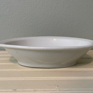 May include: A white ceramic bowl with a slightly rounded rim. The bowl is empty and sitting on a light wood surface.