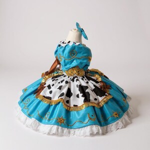 May include: A turquoise and white dress with a cow print skirt and gold trim. The dress has a matching blue bow and is displayed on a mannequin. The dress is designed with a western theme.