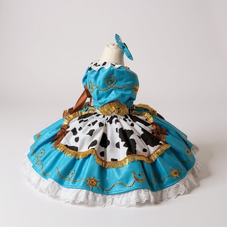 May include: A turquoise and white dress with a cow print skirt and gold trim. The dress has a matching blue bow and is displayed on a mannequin. The dress is designed with a western theme.