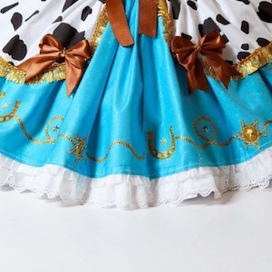 May include: A costume dress with a cow-print bodice, a turquoise skirt with gold horseshoe and star details, and white lace trim. Brown bows and gold trim add detail to this playful outfit.