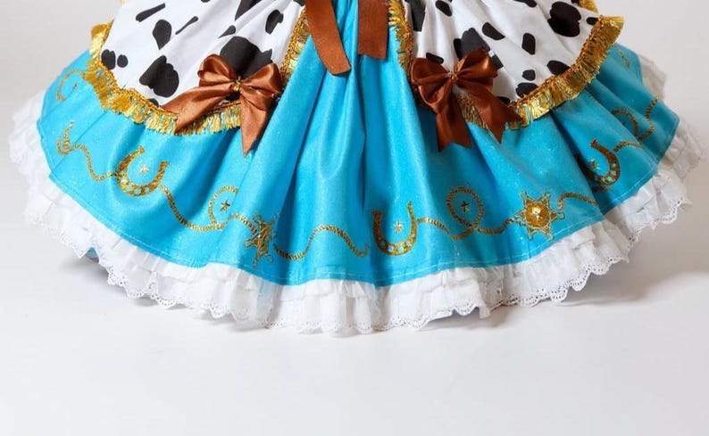 May include: A costume dress with a cow-print bodice, a turquoise skirt with gold horseshoe and star details, and white lace trim. Brown bows and gold trim add detail to this playful outfit.