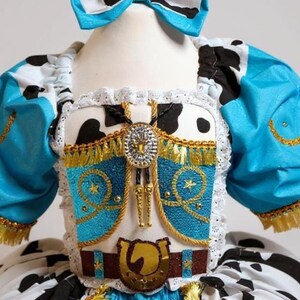May include: Cowgirl-themed costume with a blue and white cow print bow, bodice, and skirt. The costume has a turquoise bodice with gold trim, a decorative belt with a horseshoe buckle, and a bolo tie.