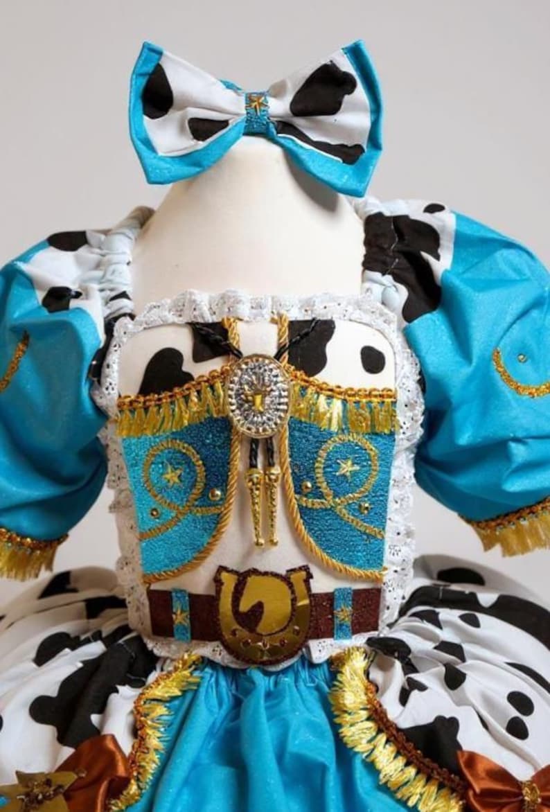 May include: Cowgirl-themed costume with a blue and white cow print bow, bodice, and skirt. The costume has a turquoise bodice with gold trim, a decorative belt with a horseshoe buckle, and a bolo tie.