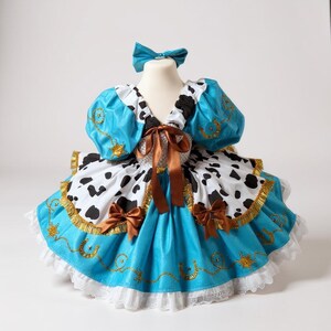 May include: A turquoise and white cowgirl-themed dress with a matching bow. The dress features a bodice with puffy sleeves, a cow-print skirt, and brown ribbon accents. Gold trim and horseshoe details add a decorative touch to this costume.