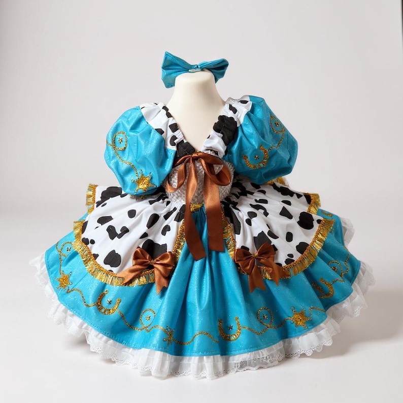 May include: A turquoise and white cowgirl-themed dress with a matching bow. The dress features a bodice with puffy sleeves, a cow-print skirt, and brown ribbon accents. Gold trim and horseshoe details add a decorative touch to this costume.