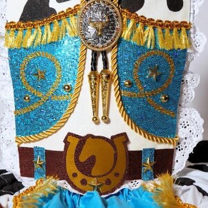 May include: A Western-style costume piece with a turquoise bodice, gold fringe, and a brown belt featuring a horseshoe and horse head. The design includes a silver and gold bolo tie with a bull emblem and star accents.