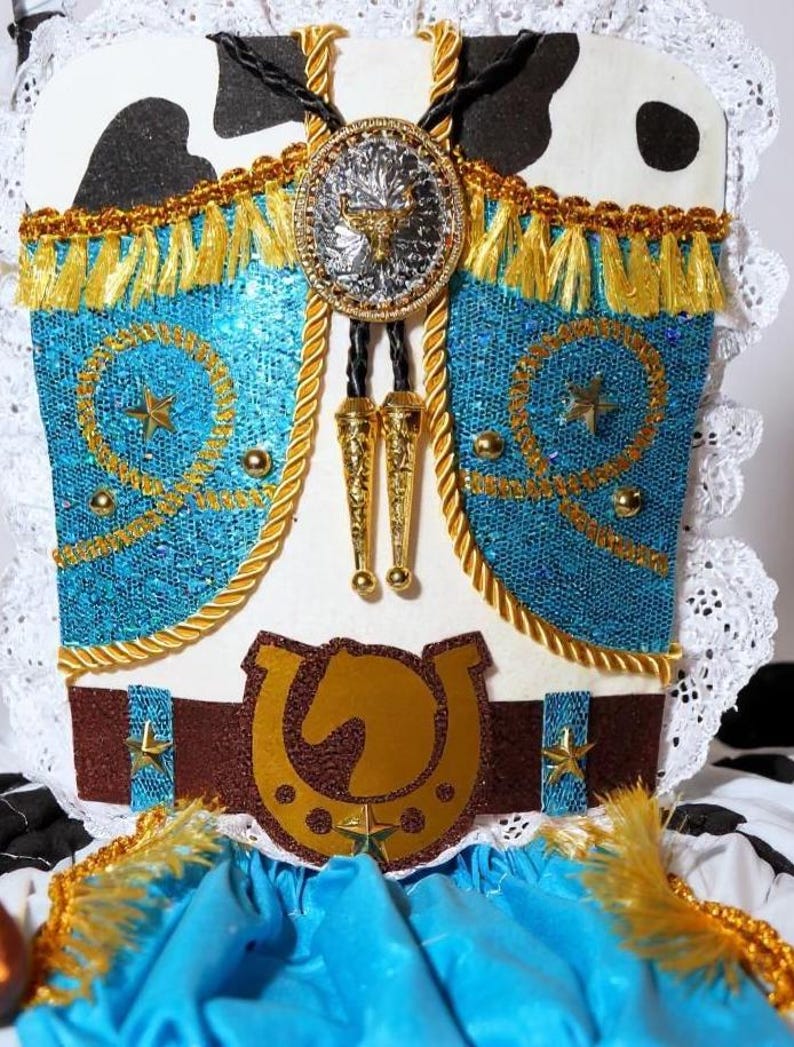 May include: A Western-style costume piece with a turquoise bodice, gold fringe, and a brown belt featuring a horseshoe and horse head. The design includes a silver and gold bolo tie with a bull emblem and star accents.