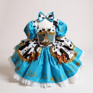 May include: A cowgirl-themed dress with a matching bow. The dress features a blue bodice and skirt, a white and black cow print skirt, and brown accents. The dress has a white lace trim and gold embellishments.