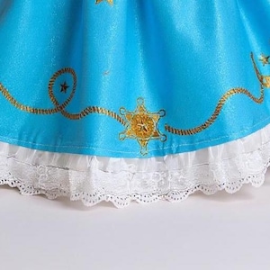 May include: A close-up of a turquoise dress with gold embellishments. The dress has a white lace trim at the hem. Gold horseshoe and star designs are printed on the turquoise fabric.