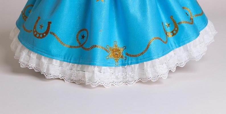 May include: A close-up of a turquoise dress with gold embellishments. The dress has a white lace trim at the hem. Gold horseshoe and star designs are printed on the turquoise fabric.