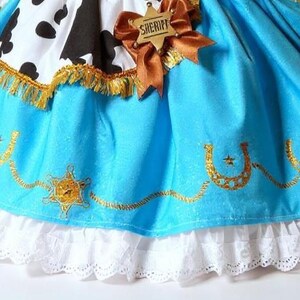 May include: A close-up of a costume skirt with a cow print top and a light blue skirt. The skirt has gold embroidery, a sheriff badge, and a white lace trim. A brown ribbon and sheriff badge are attached.