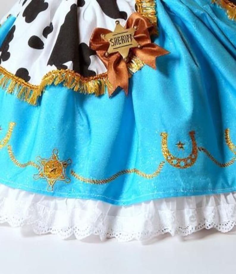 May include: A close-up of a costume skirt with a cow print top and a light blue skirt. The skirt has gold embroidery, a sheriff badge, and a white lace trim. A brown ribbon and sheriff badge are attached.