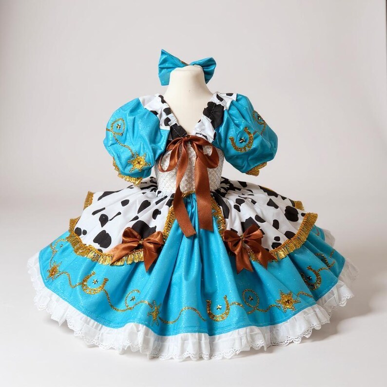 May include: A turquoise and white cowgirl-themed dress with a matching bow. The dress features a bodice with a white and black cow print, turquoise sleeves, and a turquoise skirt with gold accents and white lace trim.