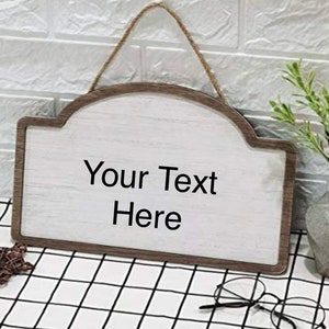 Puede incluir: Un letrero de madera blanco con un marco de madera marrón y un colgador de cuerda. El letrero tiene una parte superior redondeada y el texto "Your Text Here" está impreso en el letrero.