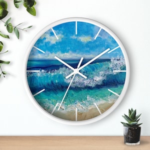Op de afbeelding: Witte ronde wandklok met een blauwe oceaan golf en wit schuim dat breekt op een zandstrand. De wijzerplaat van de klok heeft witte wijzers en zwarte cijfers.