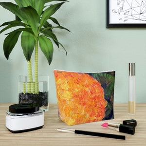 Puede incluir: Un neceser rectangular con un diseño de pintura abstracta naranja y amarilla vibrante. La bolsa es de tela y tiene una cremallera blanca. Otros artículos en la superficie de madera incluyen una planta, maquillaje y un frasco de perfume.