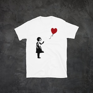 May include: White t-shirt with a black and white stencil design of a girl reaching for a red heart-shaped balloon.