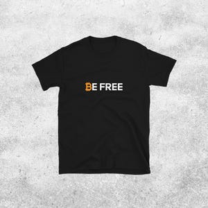 Op de afbeelding: Zwart t-shirt met de tekst "BE FREE" in witte hoofdletters. Het Bitcoin-symbool is oranje. Het shirt wordt gepresenteerd tegen een lichtgrijze achtergrond.