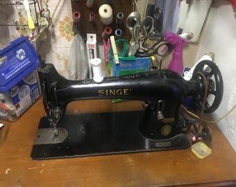 May include: A vintage black Singer sewing machine with the brand name prominently displayed. The machine sits on a wooden surface, with spools of thread and other sewing supplies visible in the background. The machine's design is classic and functional.