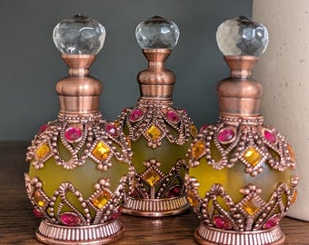 May include: Three small perfume bottles with frosted yellow glass bodies, encased in ornate copper-toned metal filigree. Each bottle features embedded pink and amber-coloured square and round rhinestones, a copper neck, and a clear faceted crystal stopper.