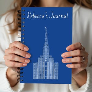 Puede incluir: Un diario azul con espiral con el texto "Rebecca's Journal" en escritura blanca. La portada presenta una ilustración arquitectónica blanca. El diario está en manos de una persona.