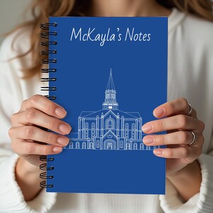 May include: A spiral-bound notebook with a blue cover featuring a white architectural illustration and the text "McKayla's Notes." The notebook's design includes a detailed drawing of a building with a steeple.