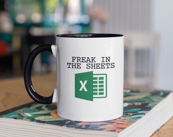 Puede incluir: Taza de cerámica blanca con asa y borde interior negros. La taza presenta el texto "FREAK IN THE SHEETS" sobre un logotipo verde de Excel. La taza está sobre un libro con un diseño floral.