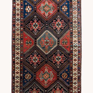 May include: A rectangular, patterned rug with a central diamond design in red, blue, and cream. The rug features geometric shapes and floral motifs, with a dark blue background and a cream border. The rug is suitable for a living room or bedroom.