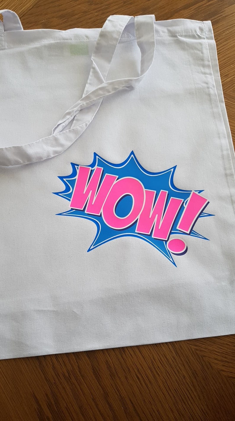 K&ouml;nnte beinhalten: Wei&szlig;e Stofftasche mit einem Comic-artigen Grafikdruck in Blau und Pink, der "WOW!" sagt.