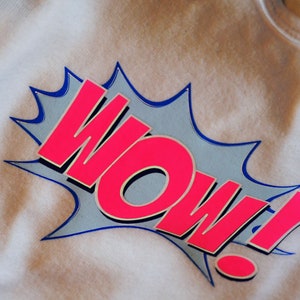 K&ouml;nnte beinhalten: Ein wei&szlig;es T-Shirt mit einer pinkfarbenen "WOW!" Grafik in einer blauen und wei&szlig;en Sprechblase im Comic-Stil.