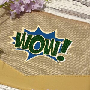 K&ouml;nnte beinhalten: Eine beige Canvas-Tasche mit Rei&szlig;verschluss und einem gr&uuml;nen und blauen Glitzer-Aufdruck "WOW!"