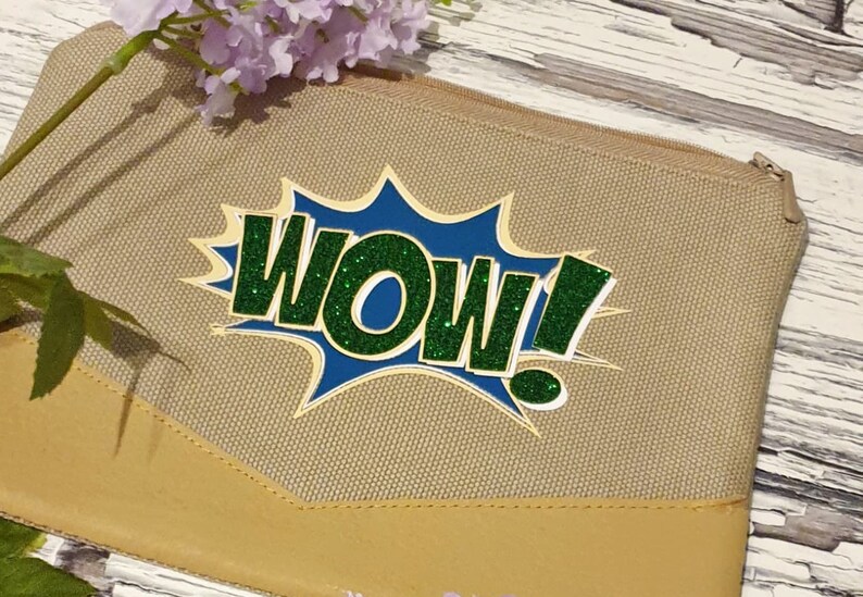 K&ouml;nnte beinhalten: Eine beige Canvas-Tasche mit Rei&szlig;verschluss und einem gr&uuml;nen und blauen Glitzer-Aufdruck "WOW!"