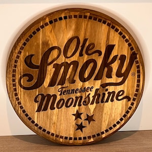 Puede incluir: Un letrero redondo de madera con una mancha marr&oacute;n. El letrero presenta el texto "Ole Smoky Tennessee Moonshine" en una fuente estilizada con un s&iacute;mbolo de marca registrada. Tres estrellas est&aacute;n debajo del texto.