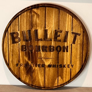 Puede incluir: Letrero redondo de madera con las palabras "BULLEIT BOURBON FRONTIER WHISKEY" talladas. El letrero está diseñado para parecerse a un fondo de barril, con listones de madera verticales y un acabado marrón oscuro.