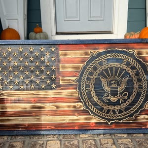 Puede incluir: Bandera americana de madera con marco azul, con estrellas y rayas talladas. El emblema de la Marina de los Estados Unidos se muestra de forma destacada en el lado derecho. La bandera es de madera y tiene un aspecto envejecido y rústico.