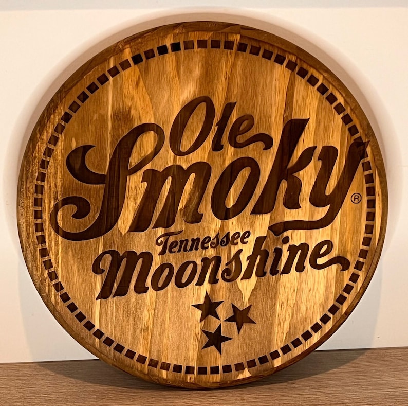 Puede incluir: Un letrero redondo de madera con una mancha marr&oacute;n. El letrero presenta el texto "Ole Smoky Tennessee Moonshine" en una fuente estilizada con un s&iacute;mbolo de marca registrada. Tres estrellas est&aacute;n debajo del texto.