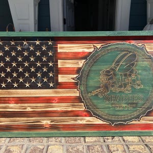 Puede incluir: Bandera americana de madera con borde verde. La bandera presenta estrellas y rayas talladas, con un emblema circular de una abeja sosteniendo una ametralladora y el texto "SEABEES" y "U.S. NAVY".