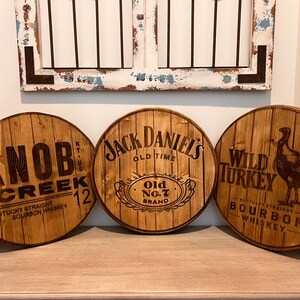 Puede incluir: Tres letreros de madera de cabeza de barril con los nombres de diferentes whiskys bourbon: Knob Creek, Jack Daniel's Old No. 7 y Wild Turkey.
