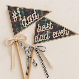 May include: Two triangular pennant flags on wooden sticks. The flags have a plaid pattern in green, red, and navy blue. One flag reads "#1 dad" and the other reads "best dad ever" in cursive. Each stick has a decorative bow.