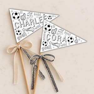 May include: Two white pennant flags on wooden sticks, decorated with soccer-themed illustrations and text. One flag says "CHARLE" and the other says "CORA". Each flag has a bow at the base of the stick. The bows are cream and black and white striped.